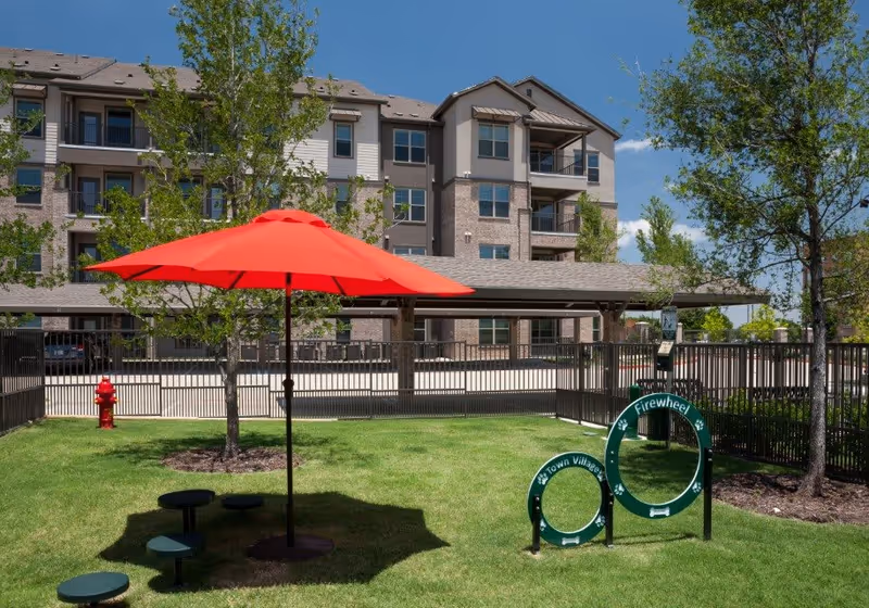 Outdoor grassy area with a large red umbrella providing shade over several small round stools. There are trees and a black metal fence surrounding the area. In the background, there is a multi-story residential building with balconies. A green circular sign with the text 'Firewheel Town Village' is visible near the right side of the image.