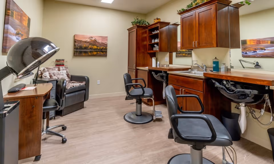 Interior of a salon room with two black salon chairs in front of sinks and wooden cabinets with mirrors. There is a black hair dryer chair on the left side, a small desk with a chair, and a black armchair with a blanket and nail polish rack next to it. The walls are decorated with landscape paintings.