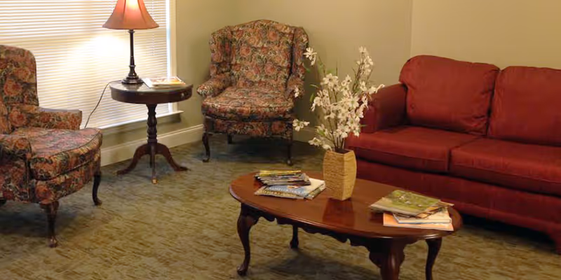 A cozy living room with a red sofa, two floral patterned armchairs, a wooden coffee table with magazines and a vase of white flowers, and a side table with a lamp next to a window with blinds.