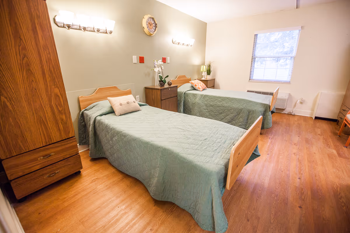 A bright bedroom with two single beds covered in green bedspreads, each with a decorative pillow. Between the beds is a wooden nightstand with a lamp and a small plant. The room has wooden flooring, a window with blinds, a wooden wardrobe, and a wall clock above the nightstand. The walls are painted in light colors, and there are two wall-mounted light fixtures above the beds.
