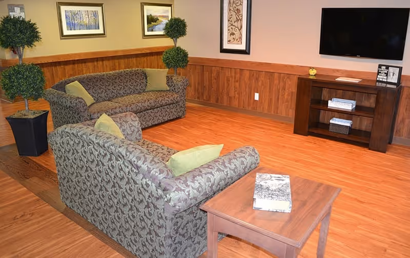 A cozy common room with two patterned sofas, an end table, mounted TV, wooden console and decorative topiary plants.