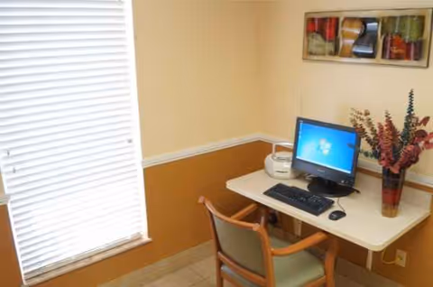 Small office desk with computer, chair, blinds, and vase against beige walls.