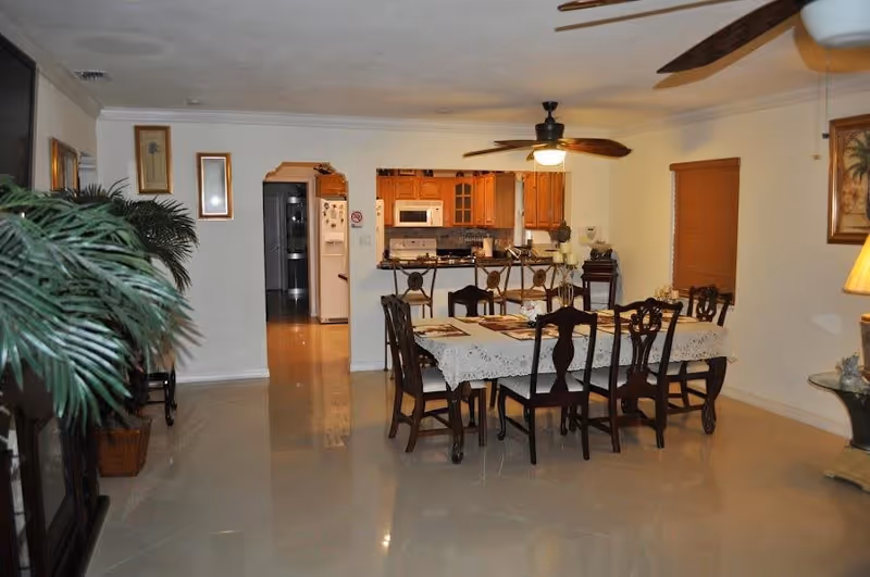 Interior view of a dining area with a rectangular table covered with a lace tablecloth and surrounded by six wooden chairs. The dining area is adjacent to a kitchen with wooden cabinets, a microwave, and a refrigerator visible through an open archway. The room has tiled floors, ceiling fans with lights, framed artwork on the walls, and a large potted plant on the left side.