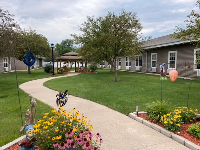 A paved walkway curves through a green lawn with flower beds containing yellow and purple flowers. Trees and decorative garden stakes are visible along the path. On the left side, there is a gazebo with seating underneath. Beige single-story buildings with white-trimmed windows surround the courtyard under a partly cloudy sky.