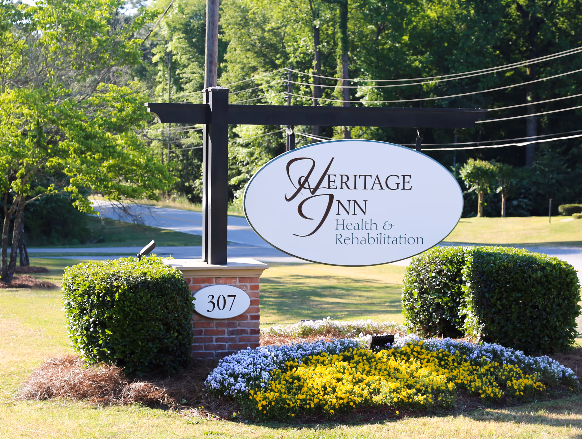 Outdoor sign for Heritage Inn Health & Rehabilitation mounted on a black post above a brick base with the number 307. The sign is surrounded by neatly trimmed bushes and a flower bed with yellow and purple flowers, with trees and a road in the background.