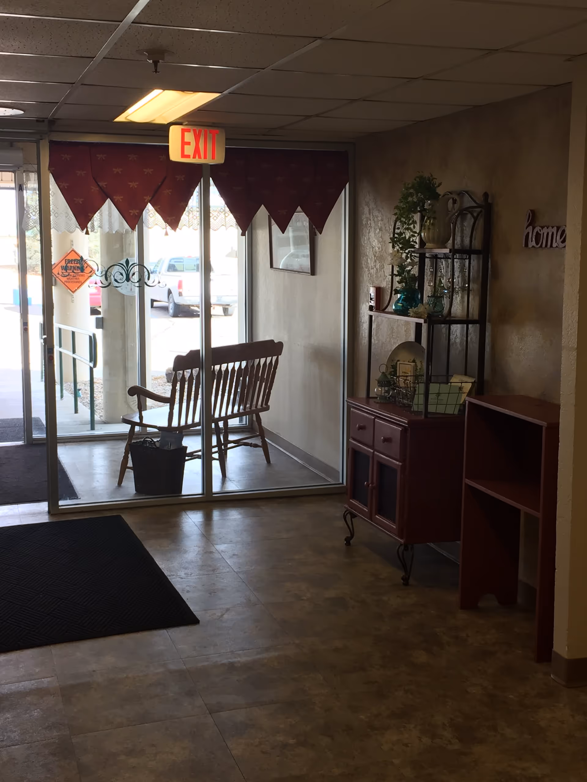 Interior view of an entryway area with a glass door leading outside. A wooden bench is visible through the glass door, and there is an exit sign hanging from the ceiling. Inside, there is a small red cabinet with decorative items and a shelf with plants and other decor. The floor has a tiled pattern and there is a black mat near the door.