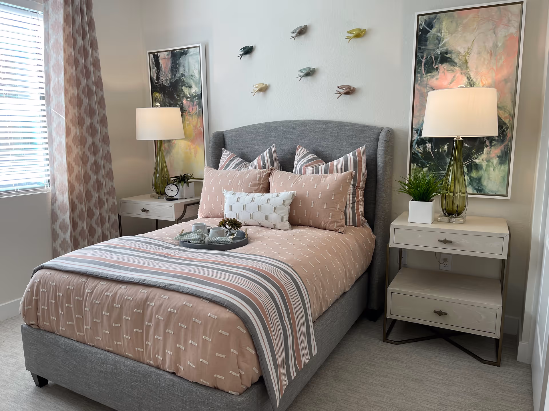 A neatly made bed with a gray upholstered headboard, pink and striped bedding, and multiple pillows in a bedroom. There are two white nightstands on either side of the bed, each with a green glass lamp and decorative items. Two abstract paintings hang on the wall behind the nightstands, and six small bird sculptures are mounted on the wall above the headboard. A window with patterned curtains is on the left side of the room.