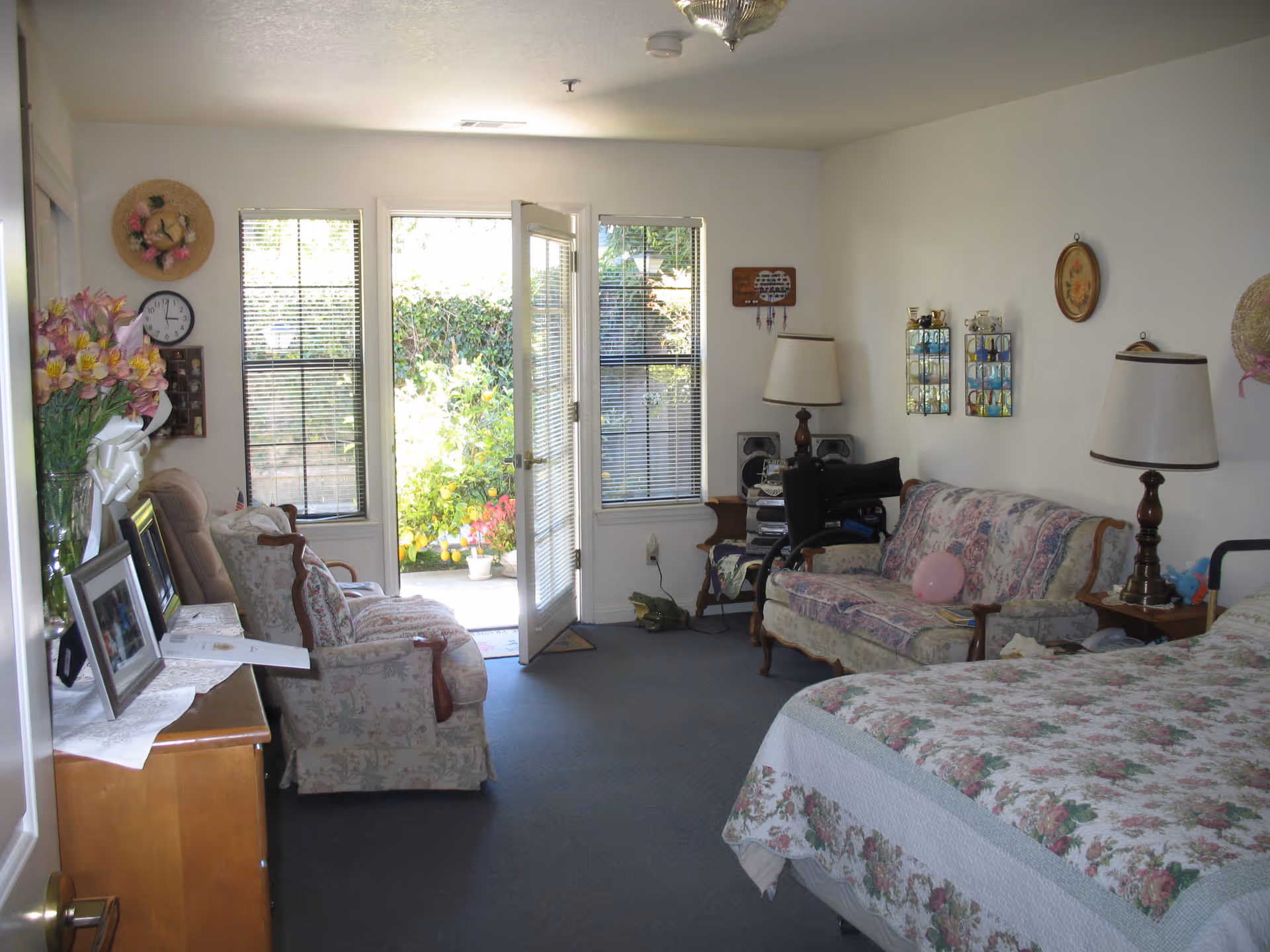 A cozy senior living room with floral-patterned furniture including a sofa and armchairs, a bed with a floral quilt, two table lamps, framed pictures on a dresser, and an open door leading to a garden area outside.