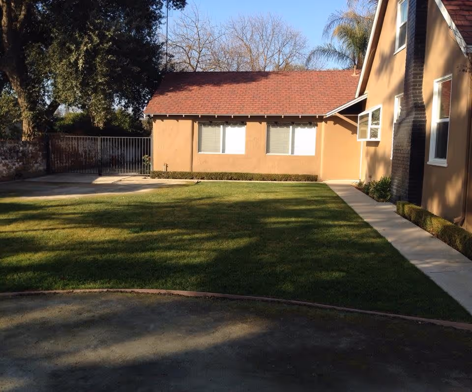 A grassy backyard area of a residential care facility with a concrete pathway along the side of a beige building with a red roof. There are trees in the background and a metal gate on the left side.