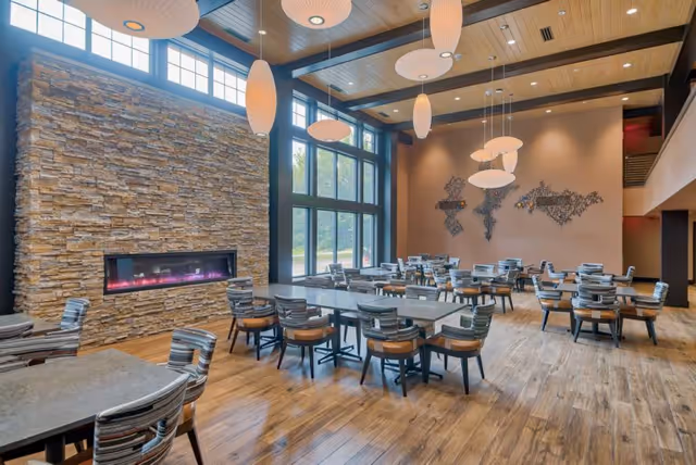 A spacious dining room with multiple tables and chairs arranged neatly on a wooden floor. The room features a large stone fireplace with a modern electric fire, tall windows letting in natural light, and contemporary pendant lights hanging from a wooden ceiling with exposed beams. A decorative wall art piece is visible on the far wall.