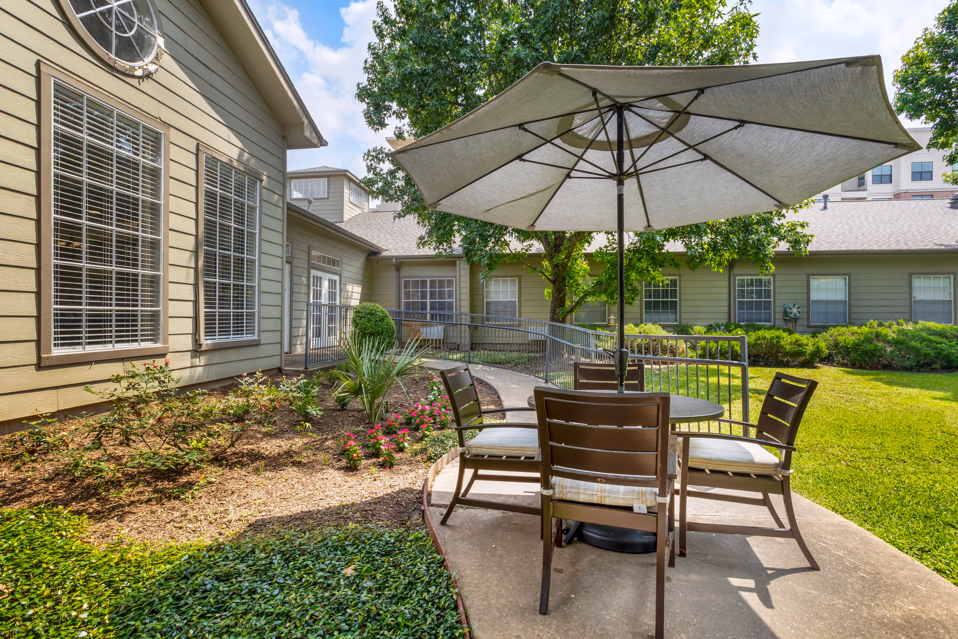 Outdoor patio area with a round table, umbrella and chairs beside a landscaped courtyard and building windows.