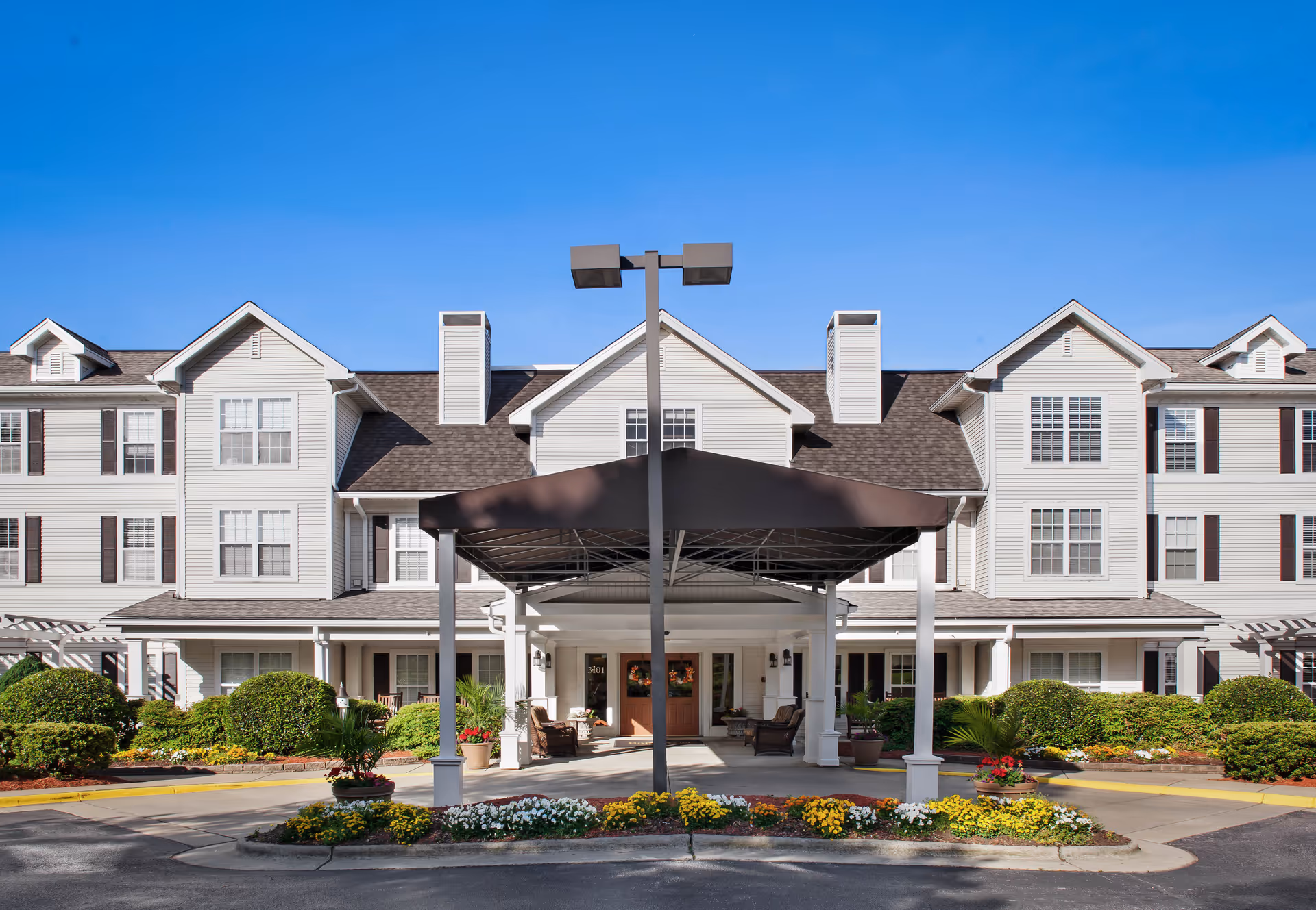 Front exterior of a three-story senior living building with a covered porte-cochere, landscaped flowerbeds, and a clear blue sky.