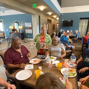 A group of elderly people sitting around a dining table in a communal dining area, eating and socializing. The room has a high ceiling, blue walls, and multiple tables with people seated. A woman in a green jacket is standing and smiling behind the table.