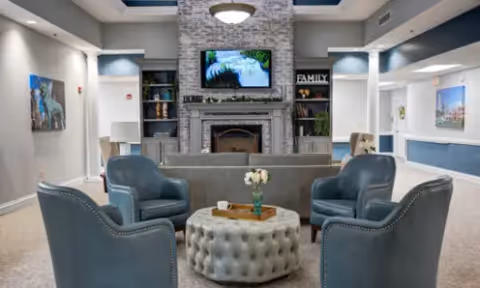 Communal lounge with blue armchairs around a tufted ottoman facing a stone fireplace and wall-mounted TV.