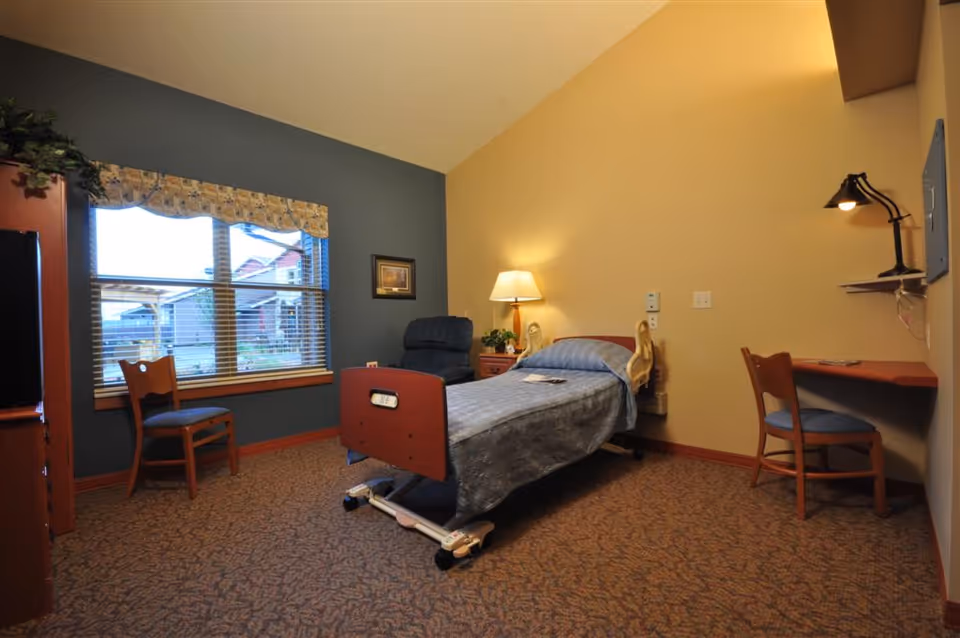 A cozy assisted living bedroom with a single hospital-style bed covered with a blue blanket, a wooden nightstand with a lamp, a blue recliner chair, two wooden chairs, a wall-mounted desk with a lamp, and a large window with blinds and a valance showing an outdoor view.