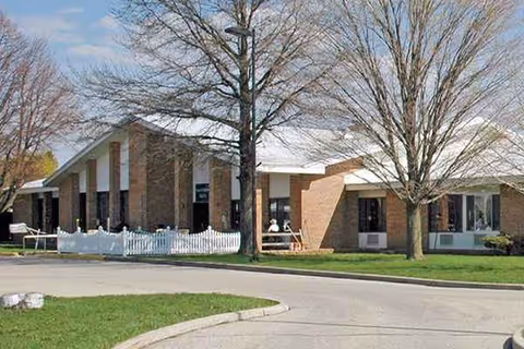 Brick exterior of a senior living facility with a white picket fence and leafless trees.