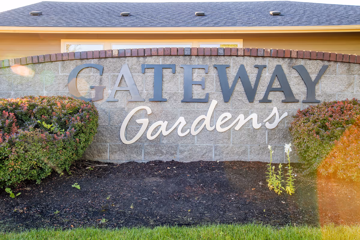 A stone and brick sign for Gateway Gardens with neatly trimmed bushes on either side and a building with a shingled roof in the background.