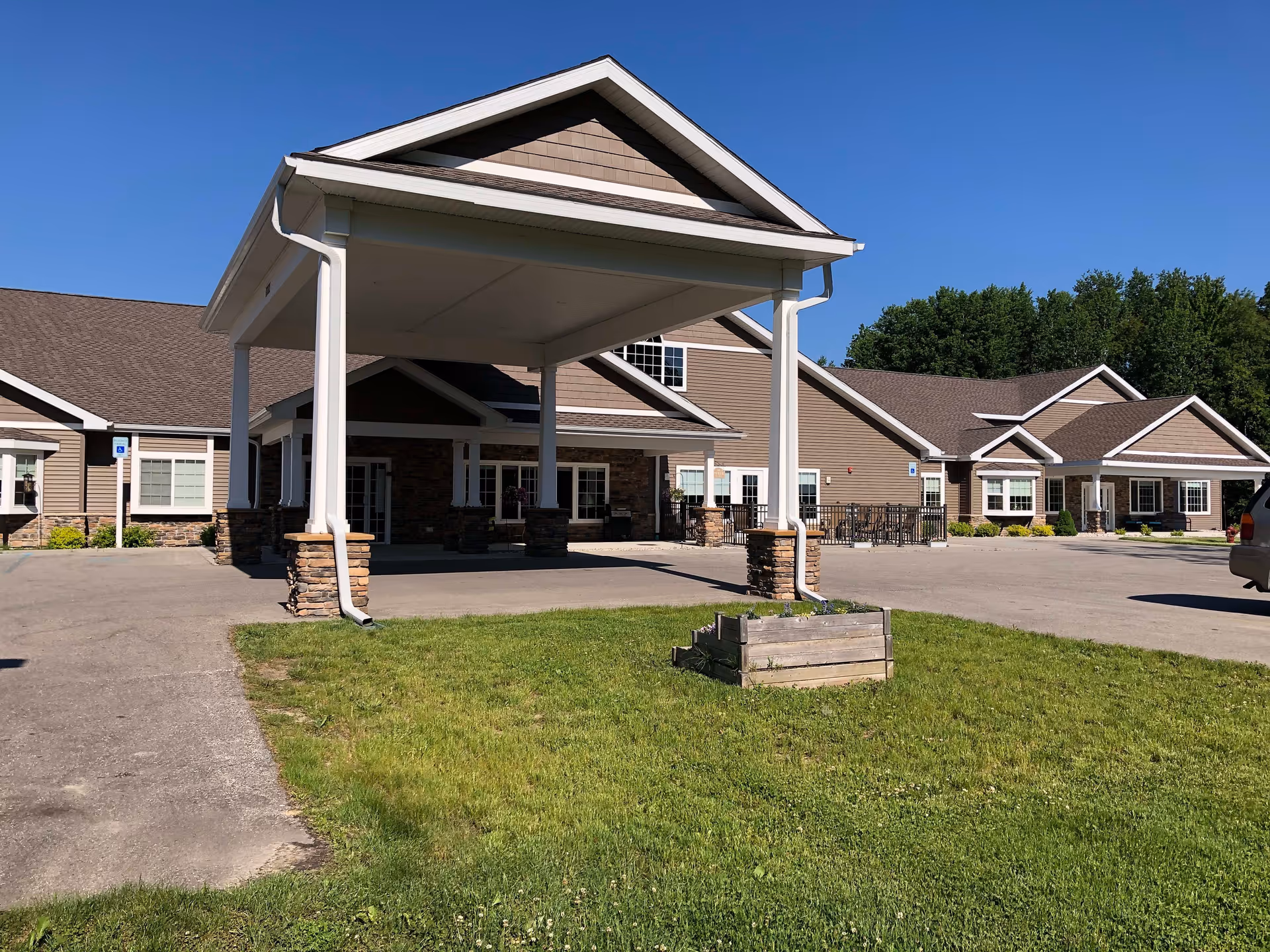 Exterior view of Reed City Fields facility showing a large covered entrance with white pillars and stone bases, a paved driveway, green lawn with a wooden planter box, and a clear blue sky.