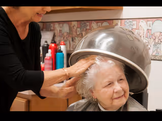 An elderly woman sitting under a hair dryer in a salon setting while a stylist is gently touching her hair, with various hair products visible in the background.