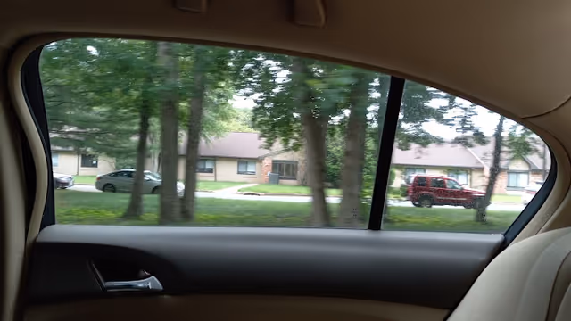 View through a car's rear side window showing trees, a grassy lawn and a single-story residential building with parked cars.