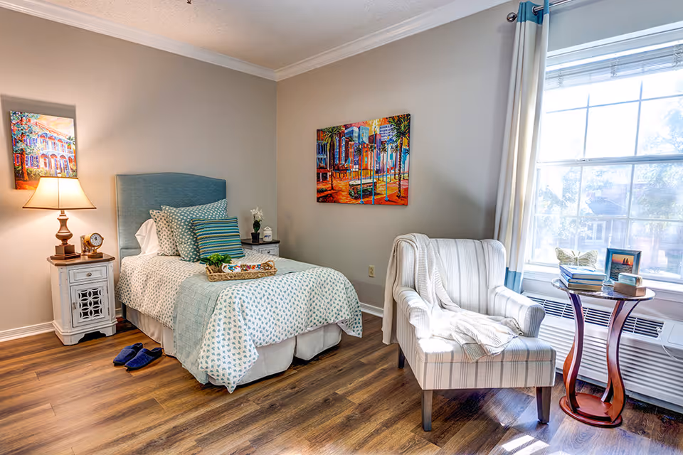 Bright single bedroom with a twin bed, patterned bedding, a bedside table and lamp, a striped armchair by a large window, and hardwood floors.