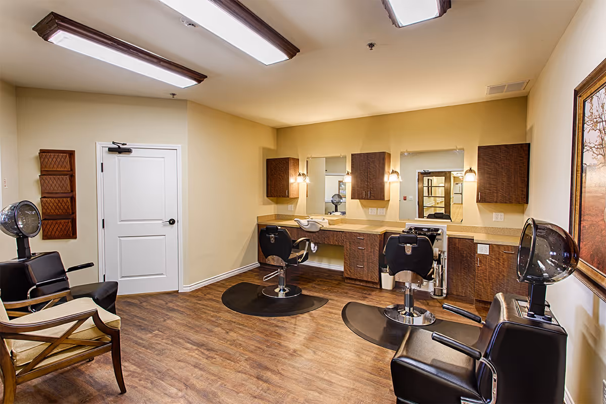 A salon-style interior with styling chairs, hooded hair dryers, mirrors, and cabinetry in a senior living facility.