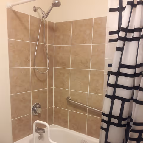 Bathtub and shower with beige tiled surround, handheld showerhead, wall-mounted grab bar, and a black-and-white patterned shower curtain.