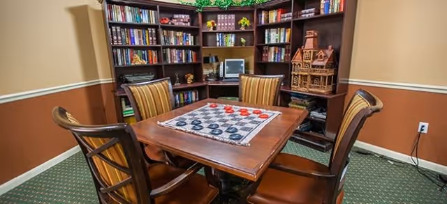 A cozy library/game room with a square wooden table, four striped chairs, a checkers board, and bookshelves lining the walls.