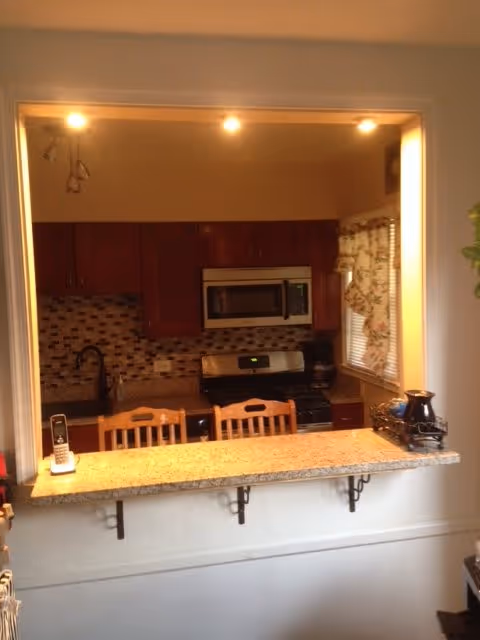 View through a pass-through into a kitchen with a granite breakfast bar and three wooden stools, stainless steel microwave and stove, and a mosaic tile backsplash.