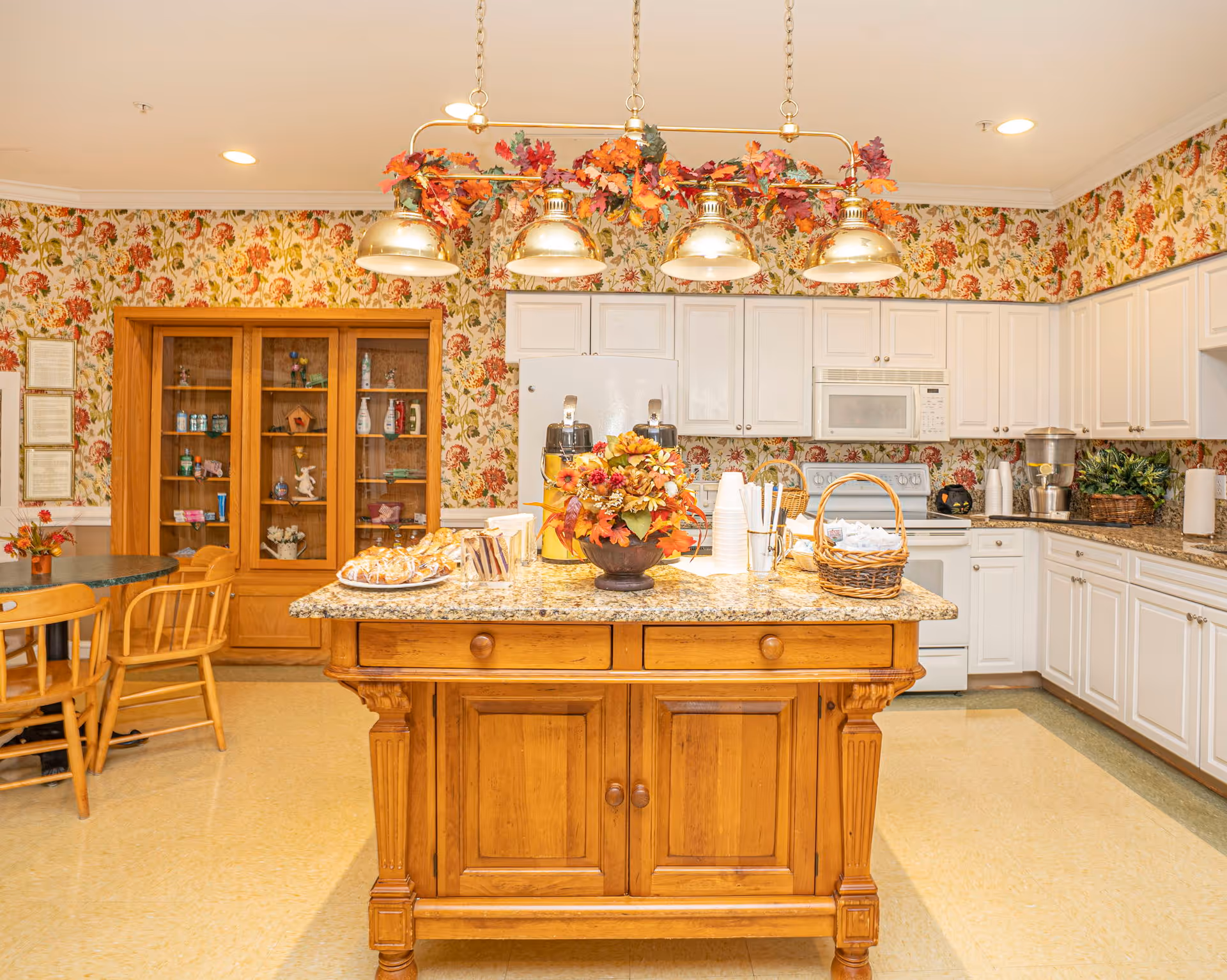 A bright kitchen area in an assisted living community featuring a central wooden island with a granite countertop decorated with a floral arrangement and baskets. The kitchen has white cabinets, a white refrigerator, microwave, and stove. There is floral wallpaper on the walls and a wooden cabinet with glass doors displaying various items. A round table with wooden chairs is visible to the left. Overhead, a light fixture with five lamps is adorned with autumn leaves.