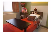 A small patient room featuring a hospital-style bed with side rails, an overbed table, stuffed animals on the bed and shelf, and a window with curtains.