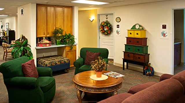 Comfortable lounge with green armchairs and a maroon sofa around a round wooden coffee table, decorative cabinet and plants.
