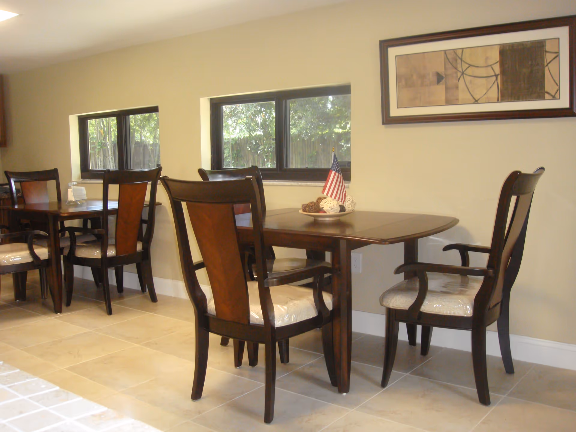 Dining area with wooden tables and chairs beside low windows and a framed artwork on the wall.