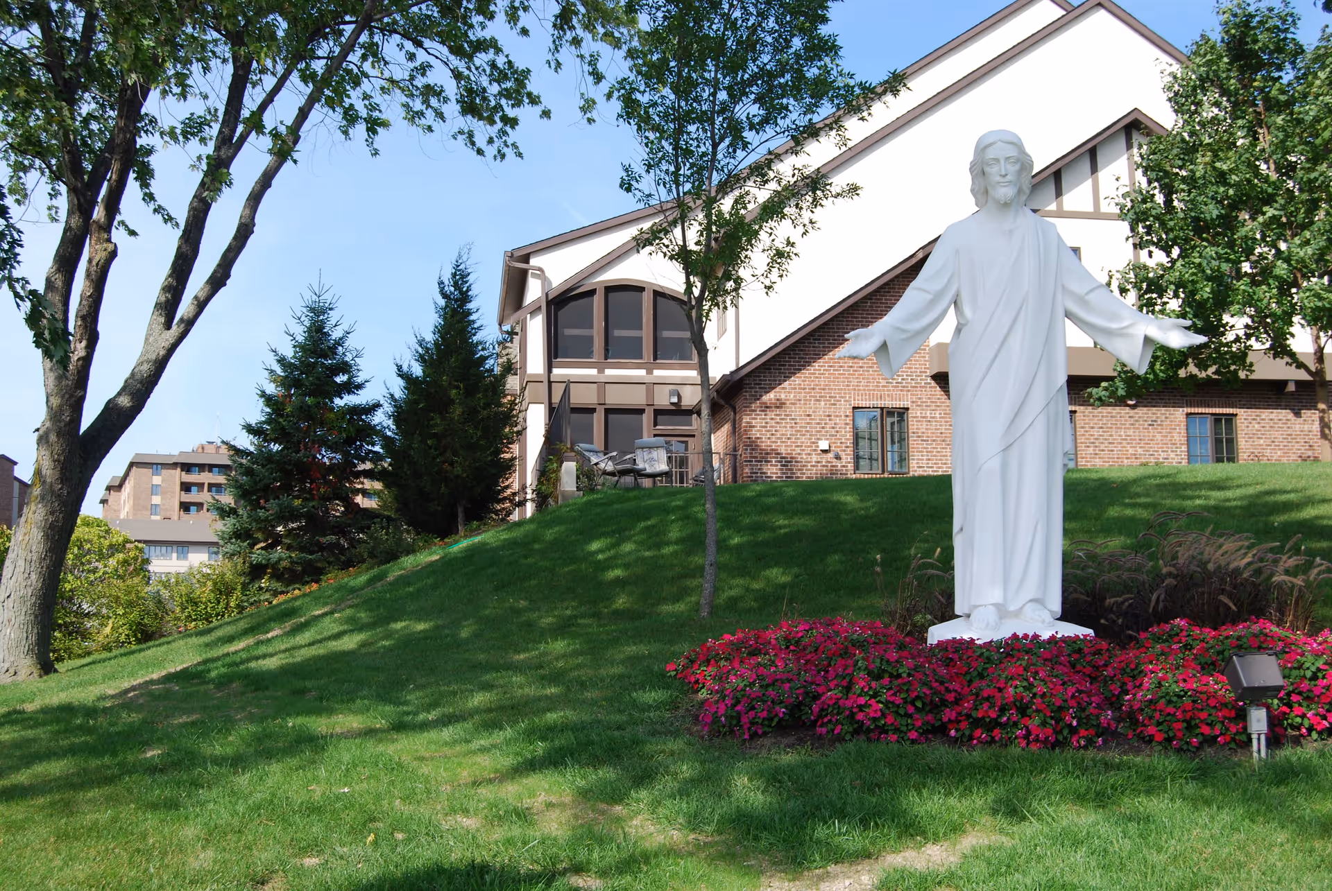 White statue of Jesus with outstretched arms on a grassy hill in front of a brick building surrounded by flowers and trees.
