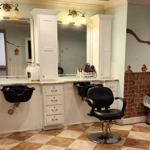 A salon corner with a black styling chair, two black shampoo bowls, mirrors, and white cabinetry under wall lights.