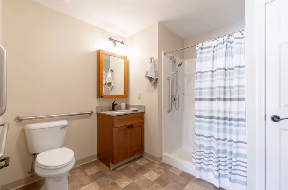 A clean, accessible bathroom featuring a white toilet with a grab bar on the wall beside it, a wooden vanity with a granite countertop and a mounted mirror above, a towel hanging on the wall, and a shower with a striped curtain and grab bars inside.