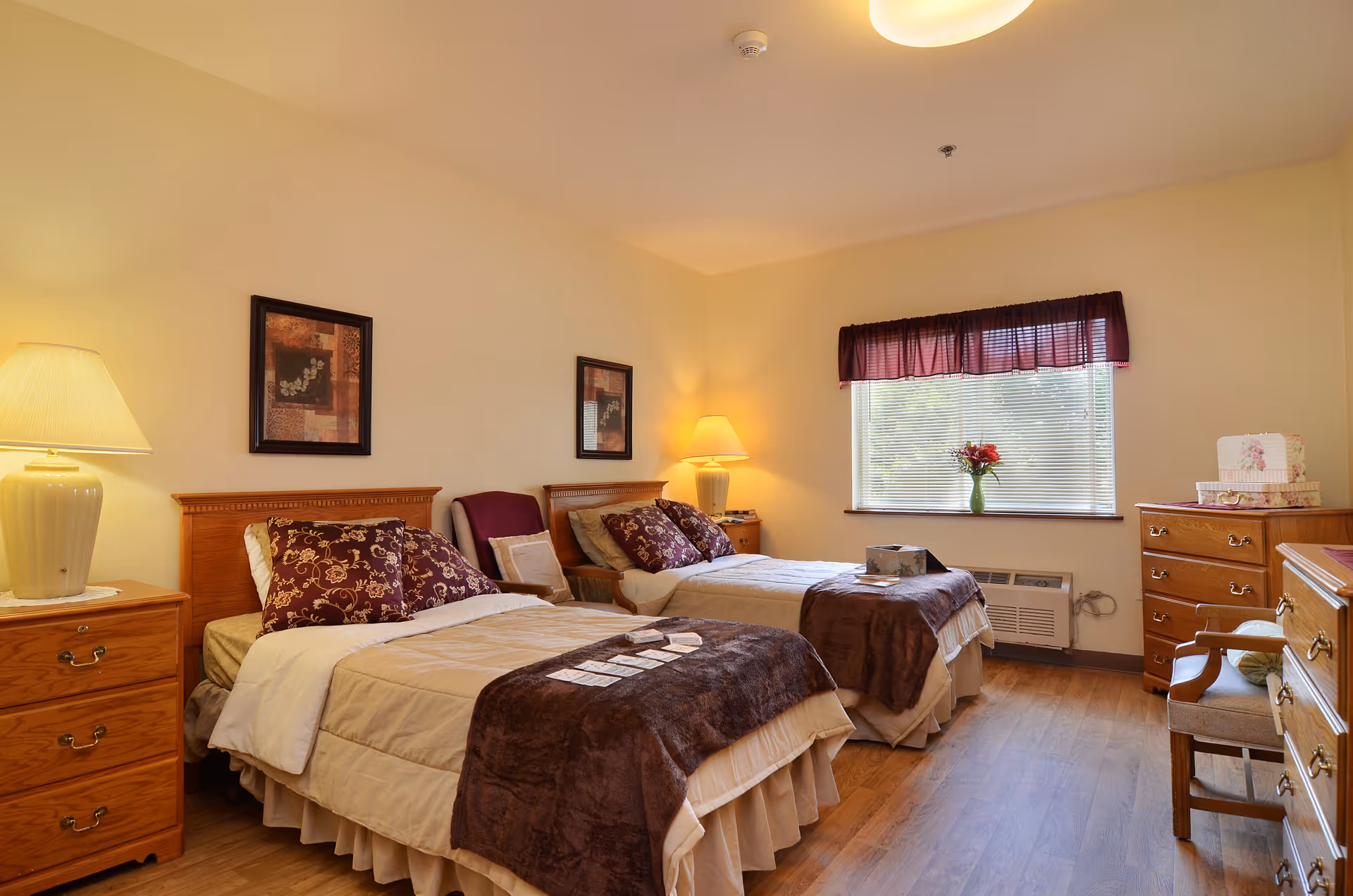A cozy bedroom with two single beds, each with beige bedding and dark brown throws. There are decorative pillows on the beds and framed artwork on the wall above the headboards. Two bedside tables with lamps flank the beds. A window with a dark valance lets in natural light, and a vase with flowers sits on the windowsill. The room also contains wooden dressers and a chair, with warm lighting and wood flooring.