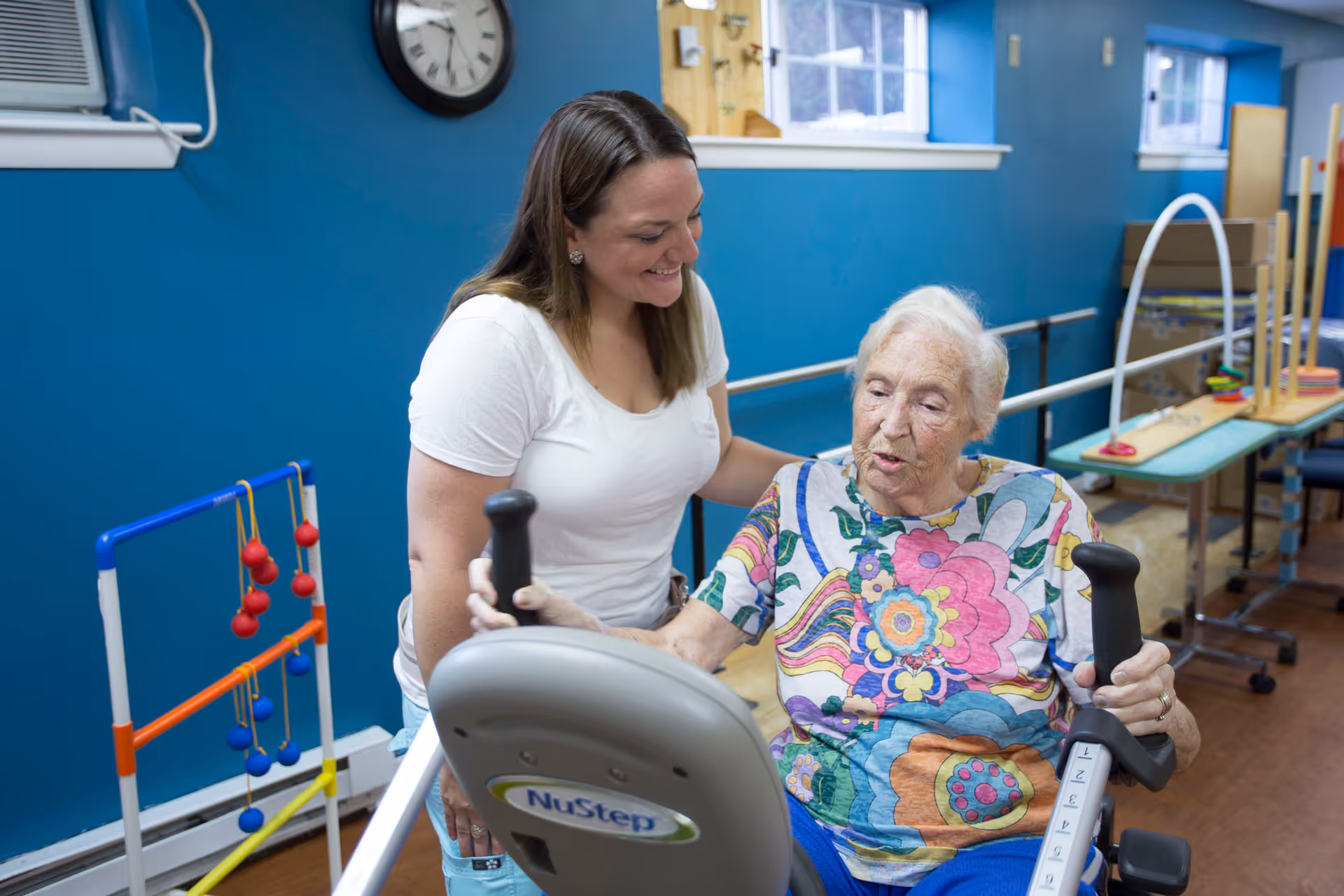 An elderly woman using a NuStep exercise machine in a room with blue walls, assisted by a smiling caregiver wearing a white shirt. The room contains various physical therapy equipment and has windows letting in natural light.