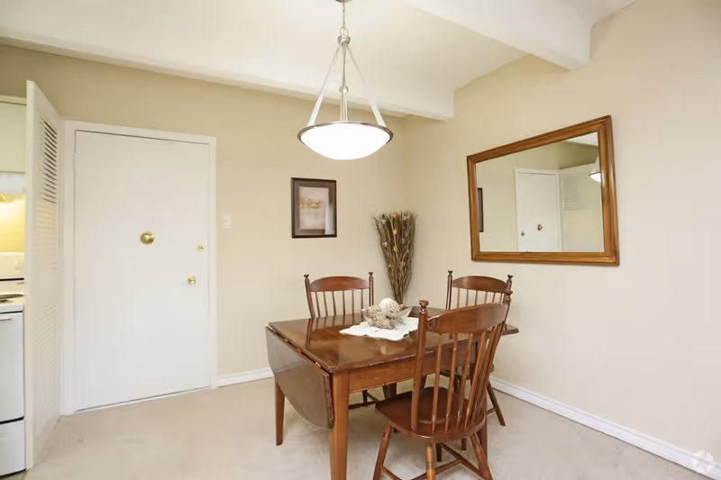 A small dining area with a wooden table and four matching wooden chairs. The table has a decorative centerpiece with a white doily and some ornamental balls. On the wall to the right is a large rectangular mirror with a wooden frame. A framed picture and a tall vase with dried plants are on the wall opposite the mirror. A white door and part of a kitchen area with a stove are visible on the left side. The room has beige walls and carpeted floor, with a hanging light fixture above the table.