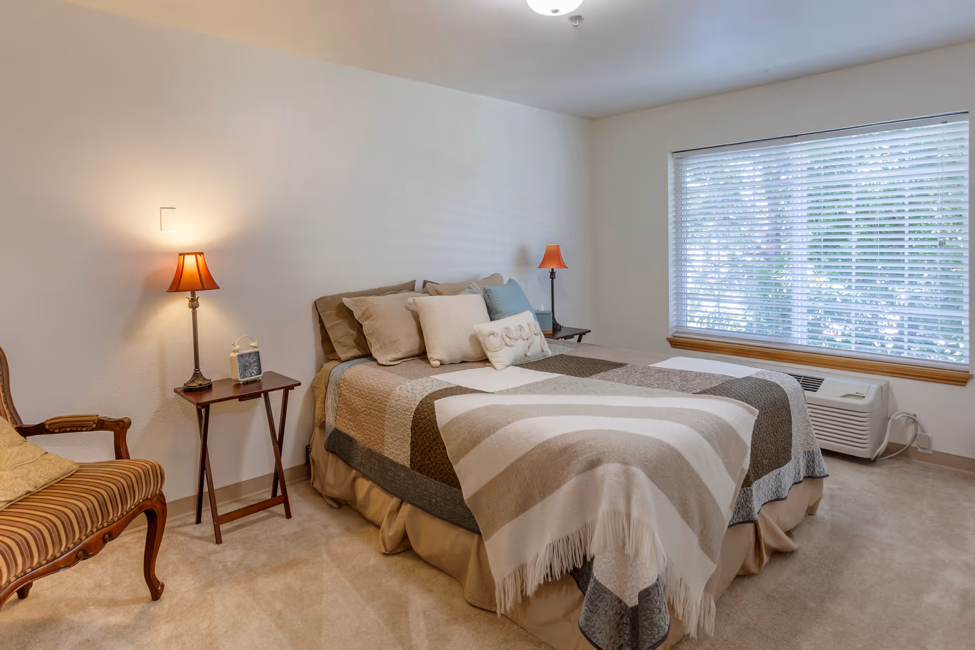 A neatly made bedroom with a bed, bedside tables and lamps, a chair, and a large window with blinds.