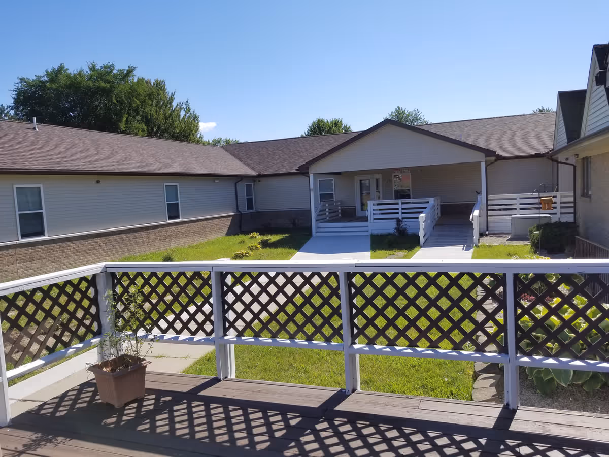 View of an outdoor courtyard area at Sandalwood Village featuring a wooden deck with lattice railing and a potted plant, green grass lawn, and a single-story building with a covered entrance and wheelchair ramp under a clear blue sky.