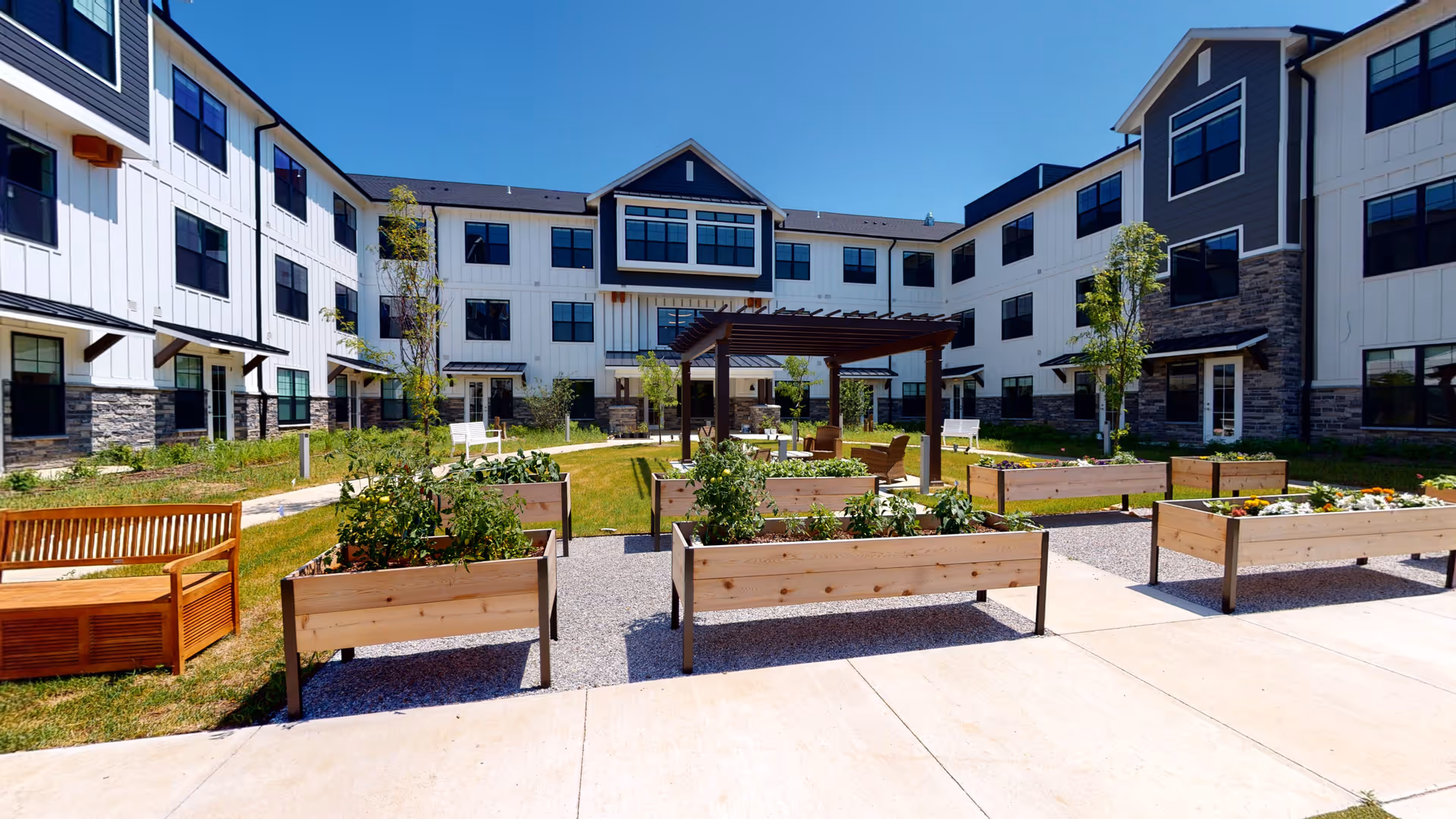 Outdoor courtyard area of a senior living facility with raised garden beds containing plants and flowers, wooden benches, a pergola with seating underneath, surrounded by a three-story building with white siding and stone accents under a clear blue sky.