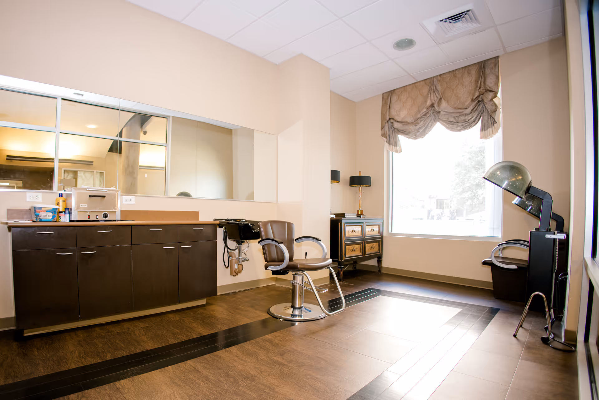 A bright room with a large window covered by a decorative valance, featuring a salon chair in front of a wall-mounted sink and a large mirror. There is a dark wooden cabinet with various items on the countertop, a vintage-style hair dryer chair, and a small chest of drawers with two lamps on top.