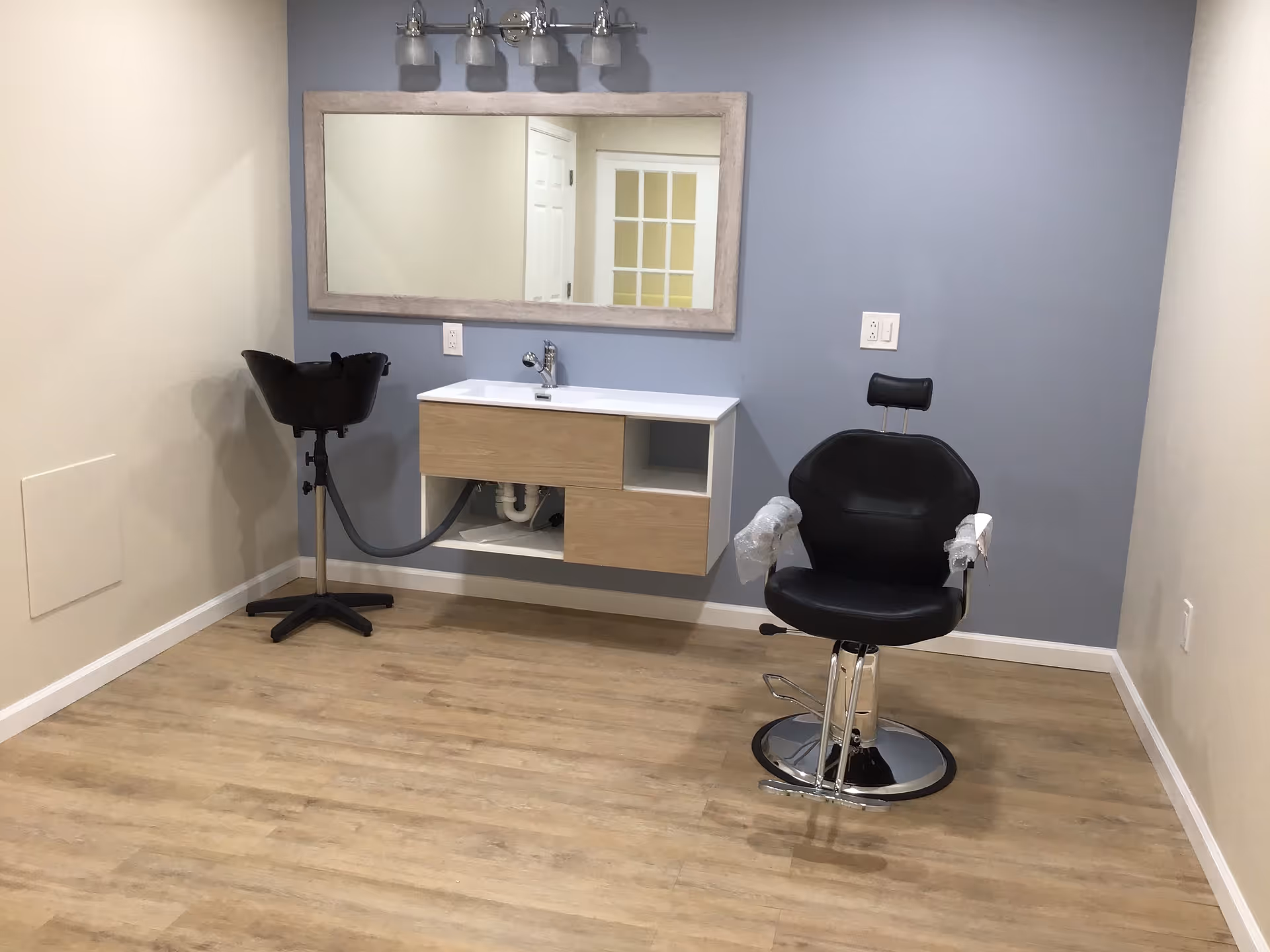 Small salon/styling room with a black barber chair, wall-mounted sink, shampoo bowl and large mirror.