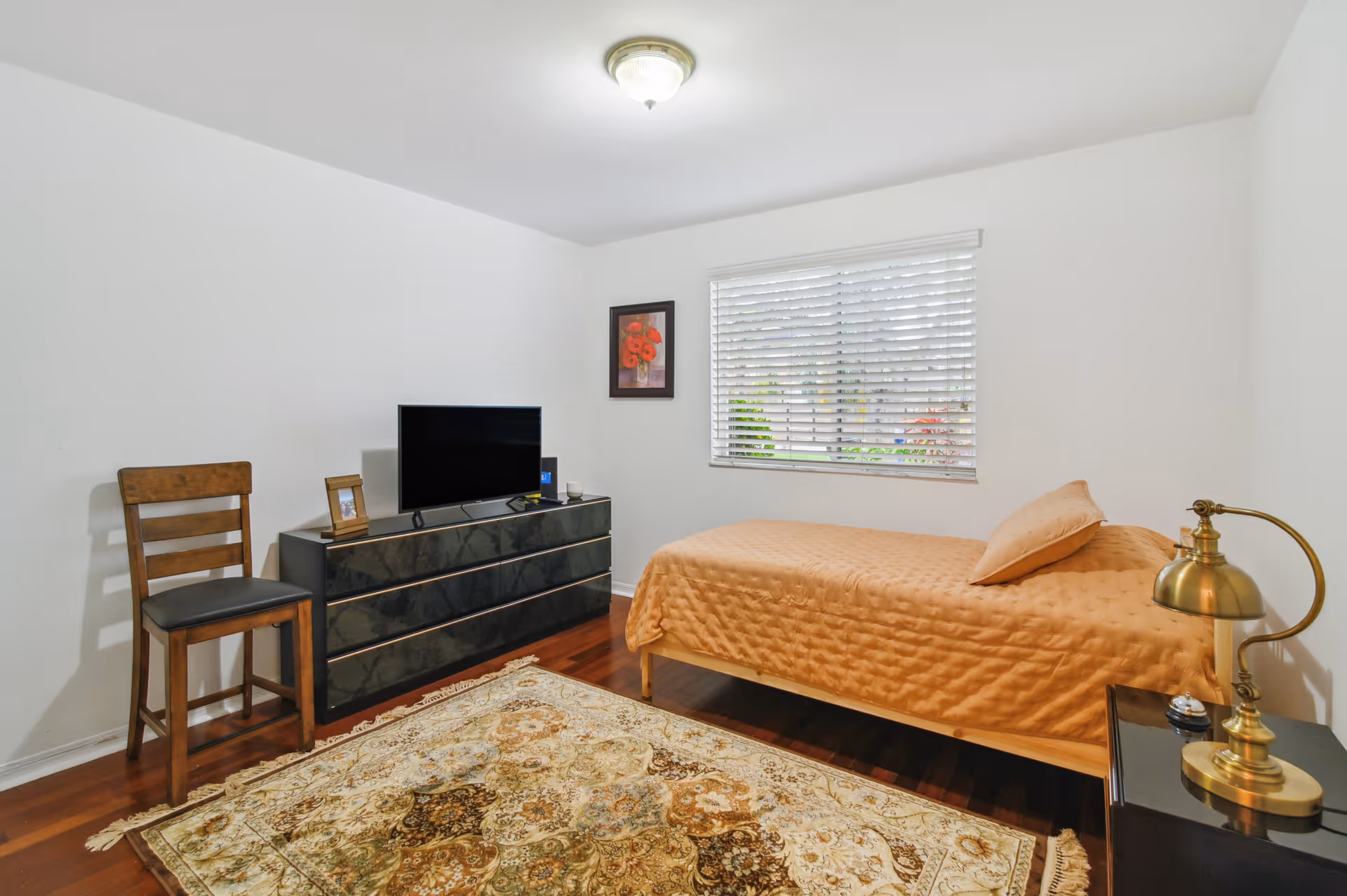 A simple bedroom with a single bed covered in a beige quilt and matching pillow. Next to the bed is a black nightstand with a brass lamp. Across from the bed is a black dresser with a flat-screen TV, a framed photo, and a small device on top. A wooden chair is placed beside the dresser. The room has white walls, a window with white blinds, and a patterned area rug on the wooden floor.
