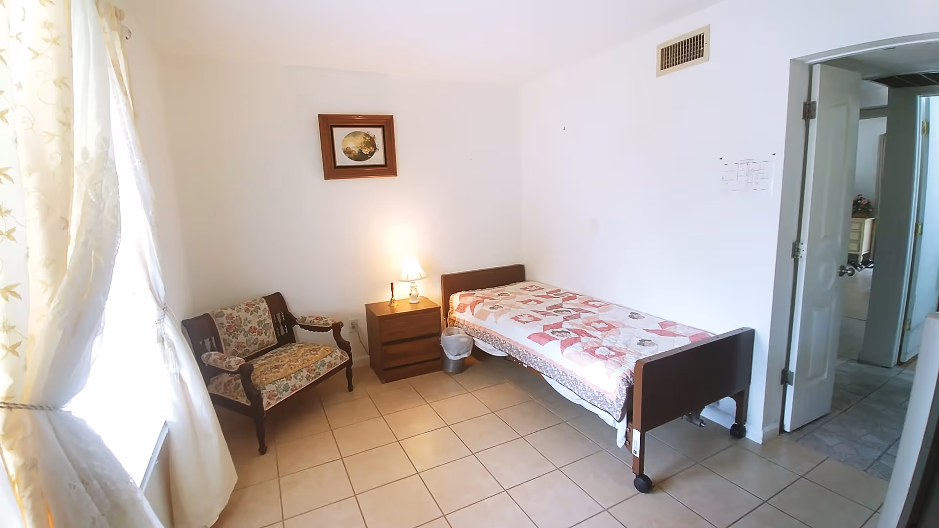 A small bedroom with a single bed covered with a patterned quilt, a wooden nightstand with a lamp and a small bell, a floral upholstered wooden chair, and a window with light-colored curtains letting in natural light. The room has tiled flooring and white walls, with a framed picture hanging above the nightstand. An open door leads to another room.