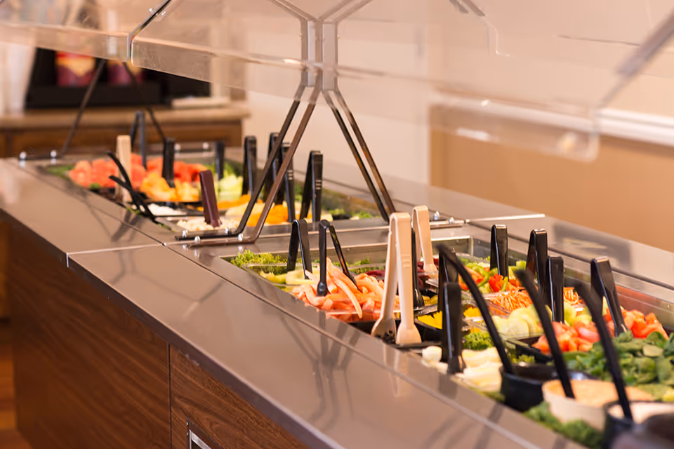 Self-serve salad bar with various vegetables and serving utensils under a clear sneeze guard.