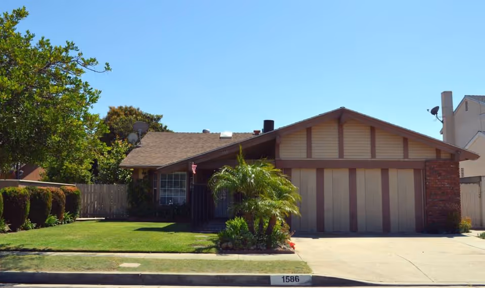 Front exterior view of a single-story residential building with a two-car garage, a well-maintained lawn, trimmed bushes, a small palm tree, and a clear blue sky.