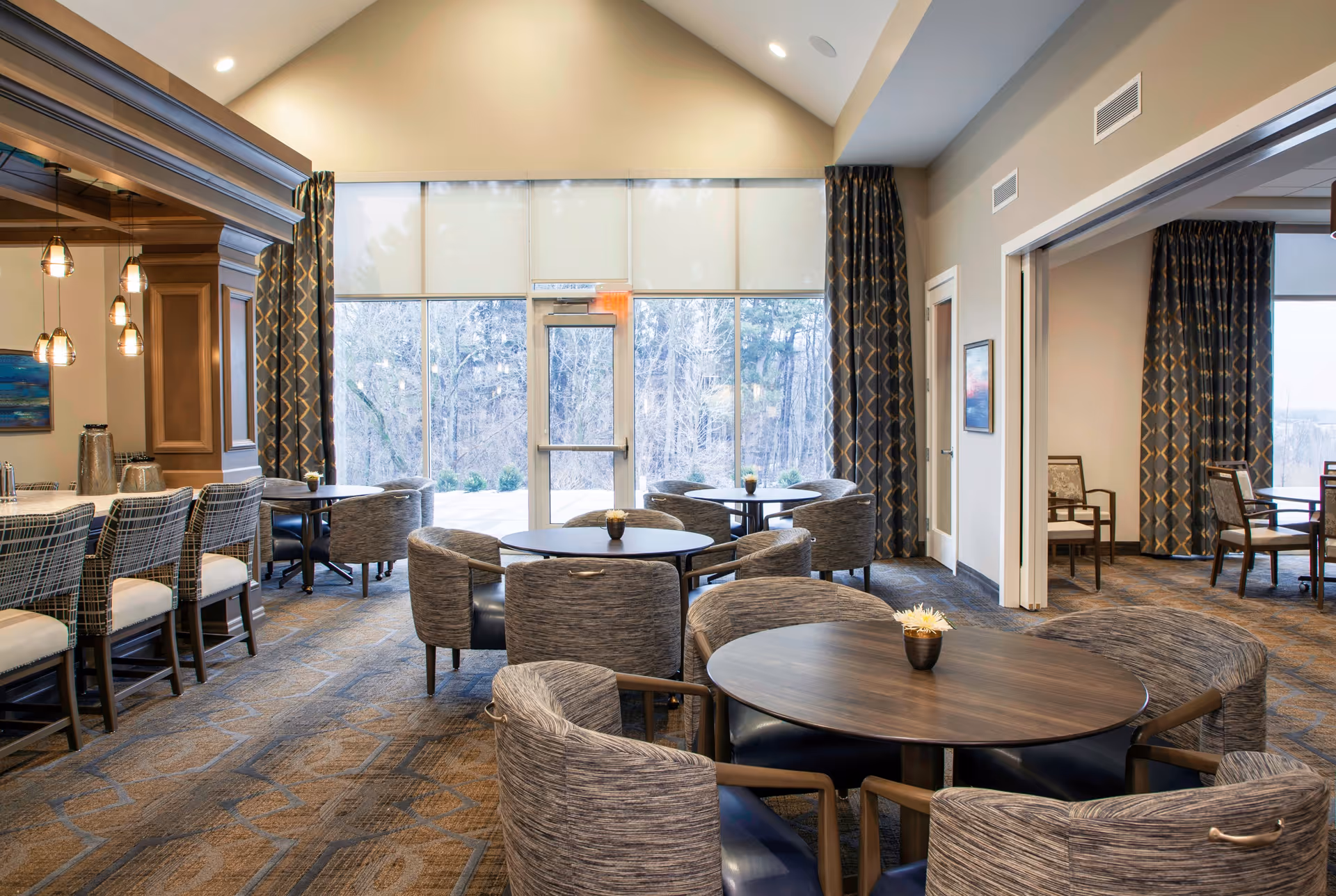 A bright and spacious dining area in Rivertown Ridge featuring round wooden tables surrounded by cushioned chairs. Large windows with patterned curtains allow natural light to fill the room, and pendant lights hang over a bar area with high stools on the left side. The room has a warm, inviting atmosphere with neutral tones and carpeted flooring.