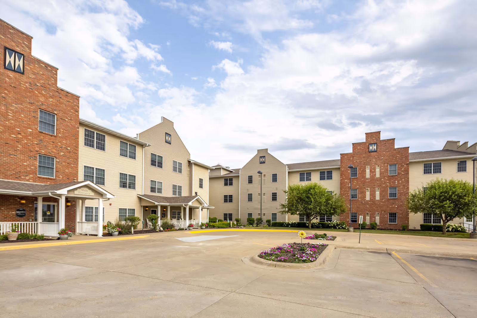 Exterior view of Independence Village of Pella, showing a multi-story senior living facility with beige siding and brick accents, a main entrance with a covered porch, landscaped flower beds, trees, and a parking area under a partly cloudy sky.
